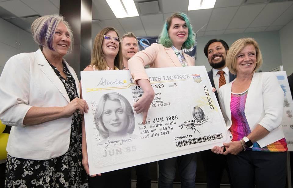 A photo of a group of people holidng up a giant version of an ID card, with Quinn Nelson pointing at the X gender marker. Around Quinn are provincial politicians, including Premier Notley